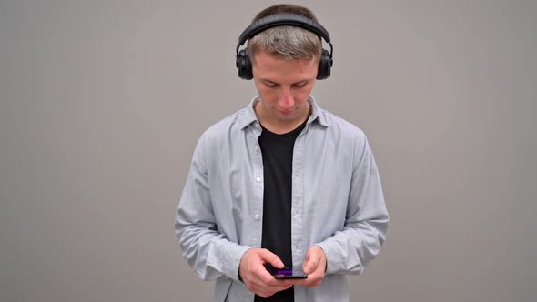 A Young Man on a Gray Background in Headphones Is Typing Text on a Smartphone