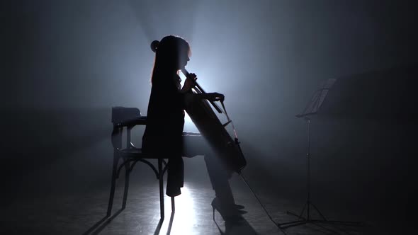 Girl Bows the Cello in a Smoky Room at Night. Silhouette. Black Smoke Background