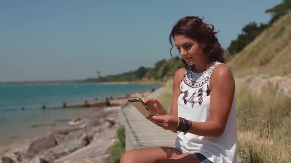 A Young Attractive Woman Sat By The Sea Makes An Online Purchase Using Her Smart Phone