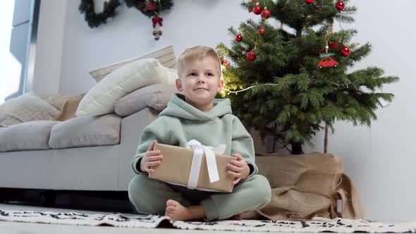 A Blond Boy in a Stylish Casual Suit Sits on the Carpet Near the Christmas Tree with a Present Box