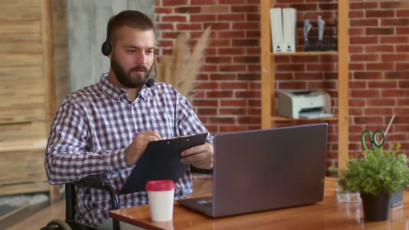 Disabled Person in Headset is Sitting with Clipboard in Front of Table with Laptop Coffee and Office