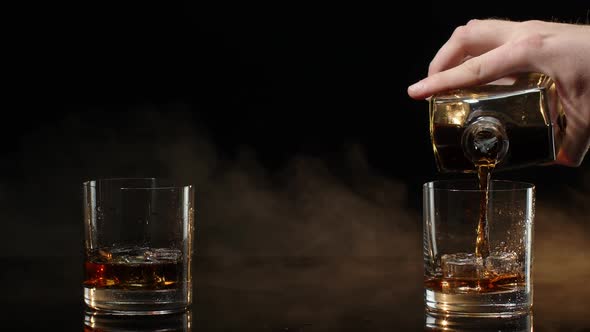 Barman Pour Golden Whiskey Cognac Brandy From Bottle Into Glasses with Ice Cubes on Dark Background