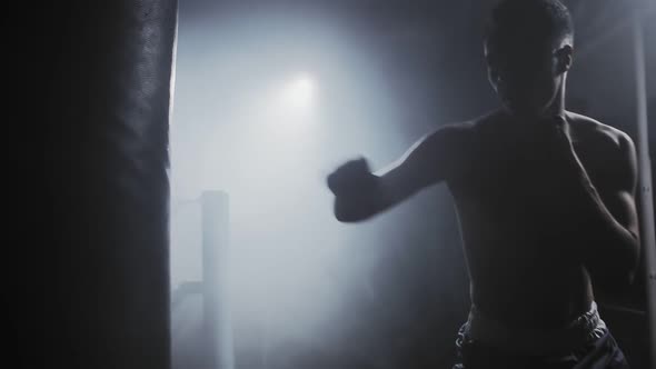 Portrait of a Caucasian male boxer beats a pear in dark gym against the background of light source,