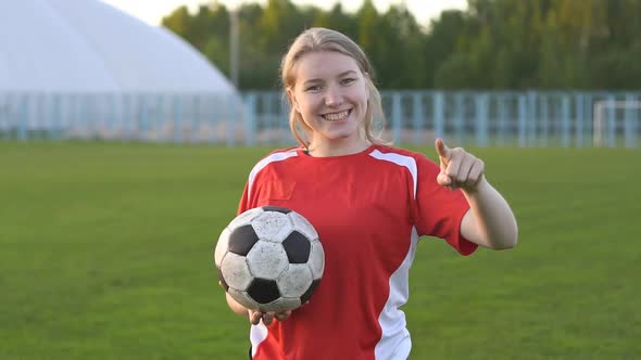 Young smiling girl football player holding soccer ball pointing with finger to the camera and to you