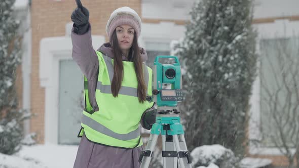 Middle Shot of Confident Young Woman Using Theodolite Outdoors on Snowy Winter Day