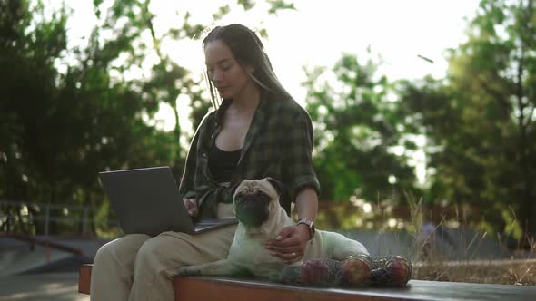 A Woman with a Laptop Sits on Parapet in a Skate Park with Her Dog Pug