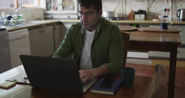 Caucasian man sitting at table, using laptop and making notes at home