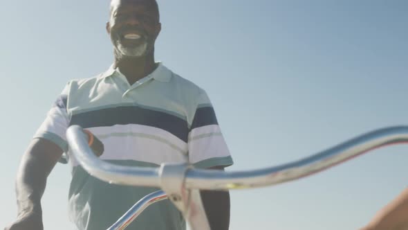 Smiling senior african american couple walking with bicycles on sunny beach