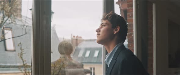 Young man standing by the window looking at the cloudy sky