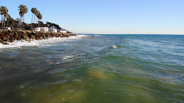 Aerial drone shot flying low over ocean waves crashing on the rocky shore with palm trees and an rv