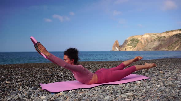 A Young Woman with Black Hair Doing Pilates with the Ring on the Yoga Mat Near the Sea on the Pebble