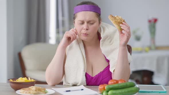 Front View of Young Obese Woman Eating Unhealthy Chips and Burger Smelling Tomato Writing Calories