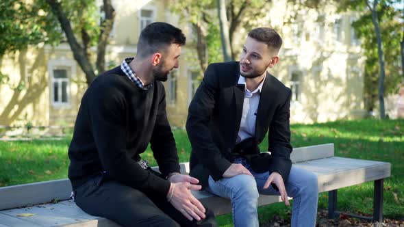 Two Confident Bearded Young Caucasian Men Talking Sitting on Bench in City Park Outdoors