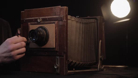 Hand of Bearded Man Wipes Lens of Old Vintage Wooden Camera on Table Under Spotlight in Isolated