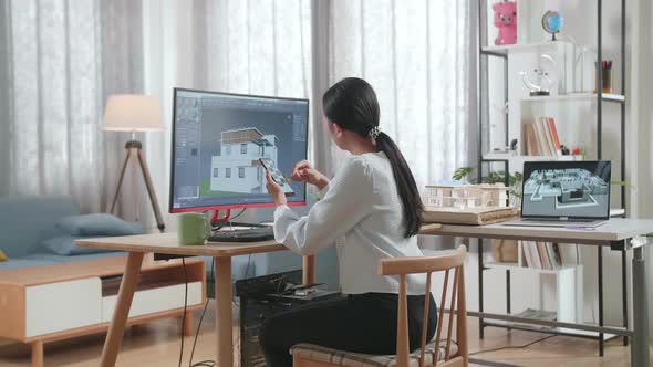 Asian Woman Engineer Using Smartphone While Designing House On A Desktop At Home