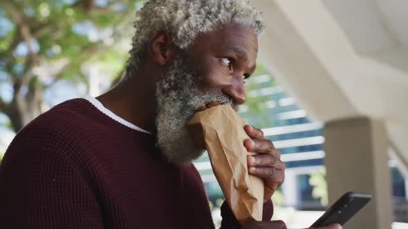 African american senior man having a snack and using smartphone at corporate park