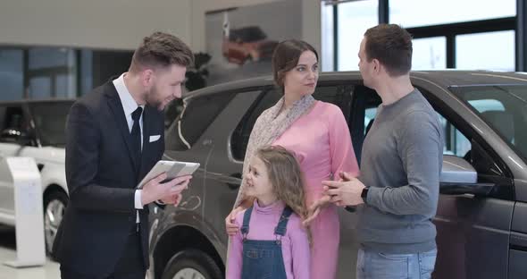 Confident Caucasian Car Dealer Presenting New Automobile To Young Family. Trader with Tablet Talking