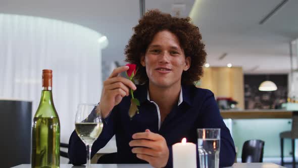 Mixed race man having romantic dinner at restaurant holding red rose and talking to camera