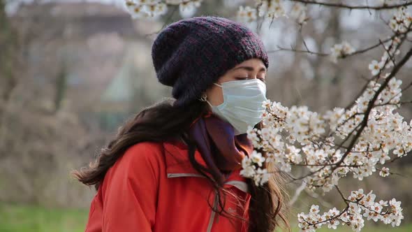 A Woman in a Medical Mask Stands on the Street Near a Flowering Tree Trying to Smell and Then