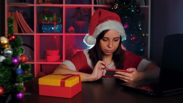 Young Woman in Santa Costume Using Smartphone in Office Sitting in Armchair in New Year's Atmosphere