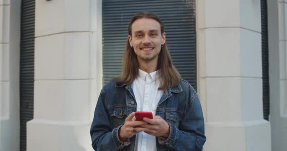 Crop View of Long Haired Guy Raising Head and Looking To Camera While Using His Smartphone. Bearded