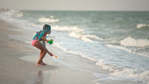 A Girl with African Braids in a Summer Costume Plays on the Beach with Shells Near the Sea with