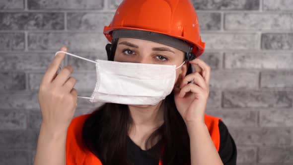 Female Construction Worker in Overalls Putting on Medical Mask on Face on Background of Gray Brick