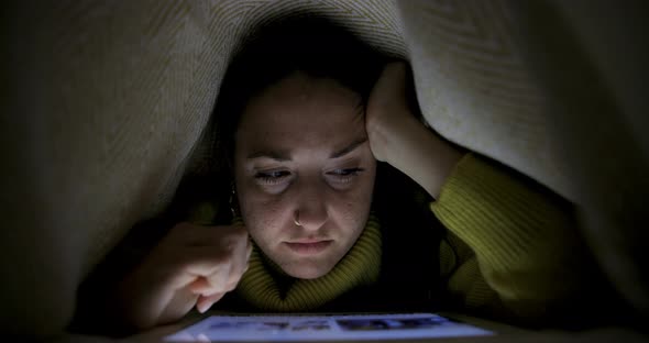 Woman on the bed under the blanket and watching photos on digital tablet