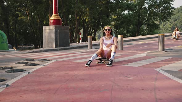 Two Young Girls with Bright Appearance Riding on a City Bridge Sitting on a Skateboard