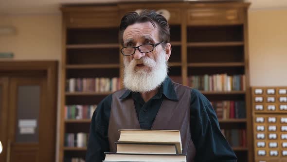 Senior Bearded Man Holding in His Hands a Lot of Books and Coming Through the Library Room