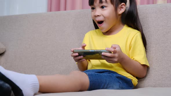 Cute little girl playing online games on her smartphone sitting on the sofa at home.