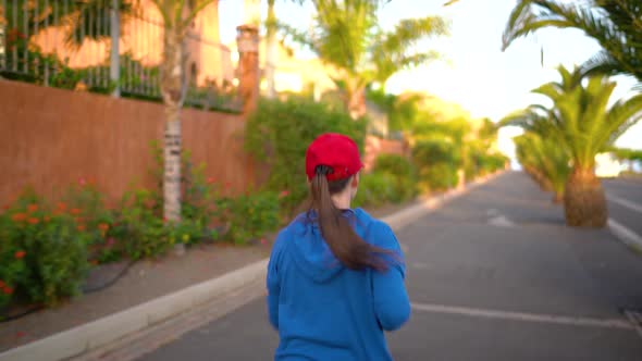 Woman Runs Down the Street Among the Palm Trees at Sunset Back View