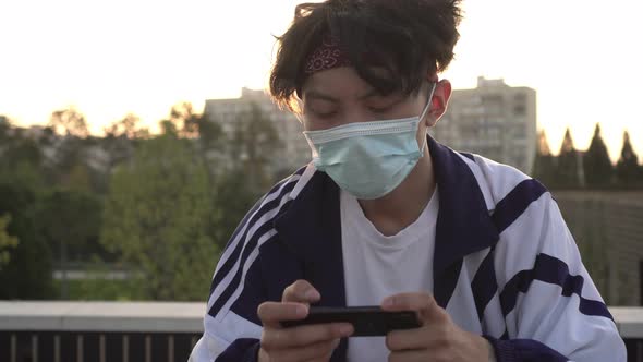 Young Asian Man in Protective Medical Mask Plays Video Games Using Smartphone Outdoors