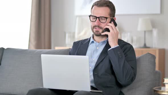 Young Businessman with Laptop Talking on Smartphone at Home 