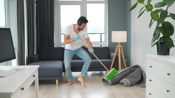 Man Listens to Music with Headphones and Cleaning the House and Having Fun Dancing with a Broom