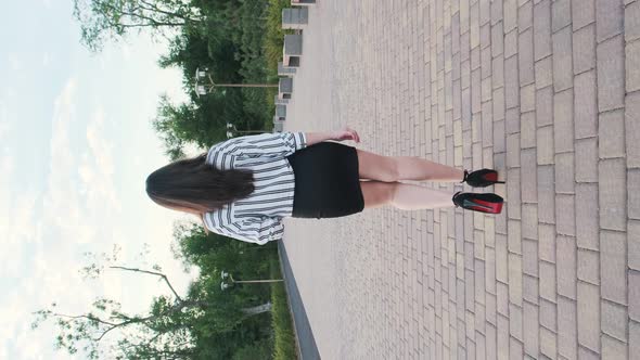 Beautiful girl in black high-heeled shoes, a black skirt and a striped shirt walks in the park. Back