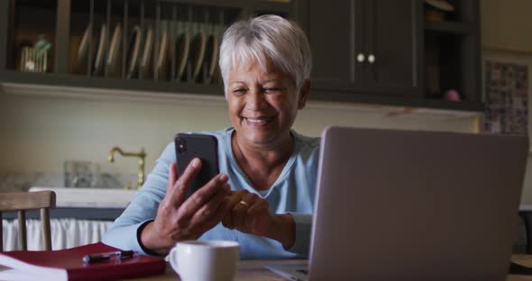 Happy senior mixed race woman using smartphone and laptop in kitchen