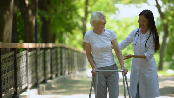 Female Doctor Helping Elderly Woman With Walking Frame, Hospital Park, Outdoors