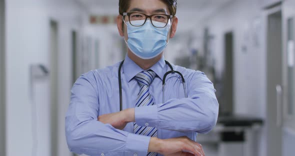 Portrait of caucasian male doctor with arms crossed wearing face mask in the corridor at hospital