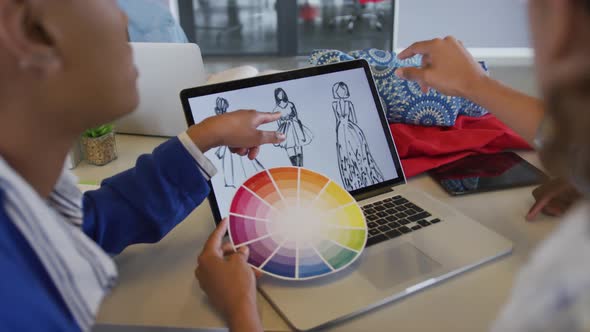 Diverse female and male designers discussing fashion designs on laptop and colour wheel in studio