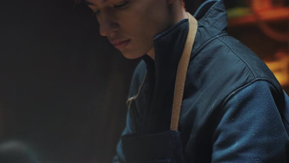 Young Attractive Carpenter is Leaning Over a Work Table in His Workshop and is Grinding Wooden