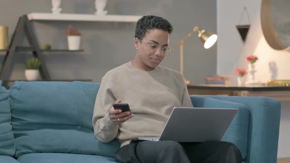 African Woman with Laptop Using Smartphone on Sofa