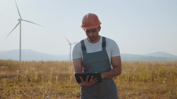 Portrait of Focused Indian Man Standing on Field with Wind Turbines and Using