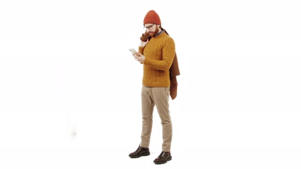Young Handsome Bearded Caucasian Man Looks at His Smartphone and Smiles Holding Coat on His Shoulder