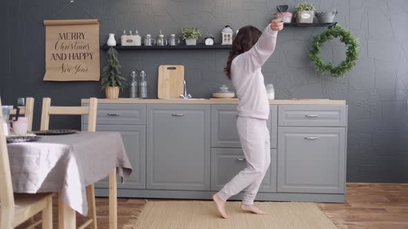 Happy Young Woman Dancing in Kitchen Wearing Pajamas in the Morning Listening To Music and Coffee at