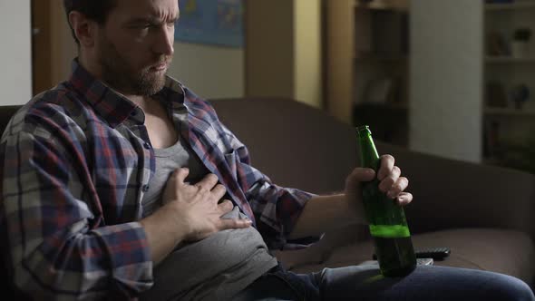 Alcohol addict scratching belly with bottle of beer in hand, dozing off after