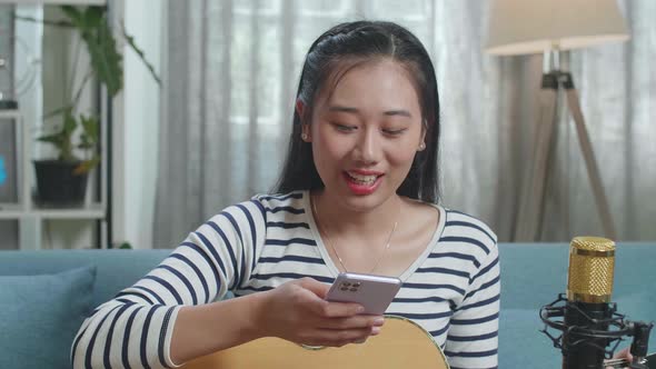 Close Up Of Asian Woman With Microphone Using Smartphone And Speaking While Playing A Guitar At Home