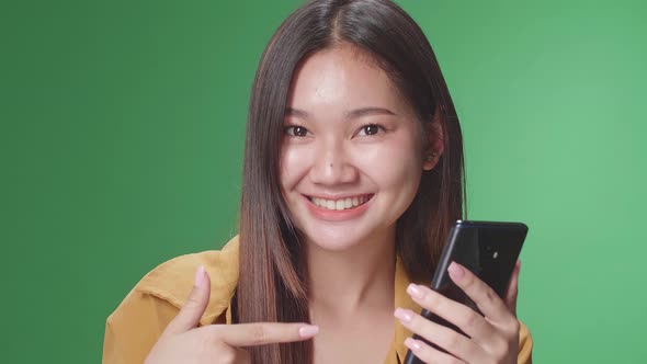 Asian Woman Use Phone And Pointing To Smart Phone While Standing In The Green Screen Studio