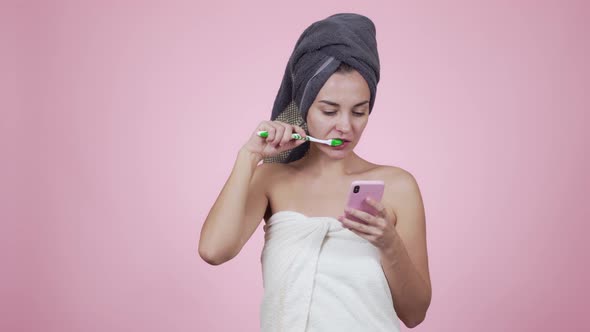 Girl in Towel Brushes Her Teeth and Uses Smartphone Isolated on Pink Background