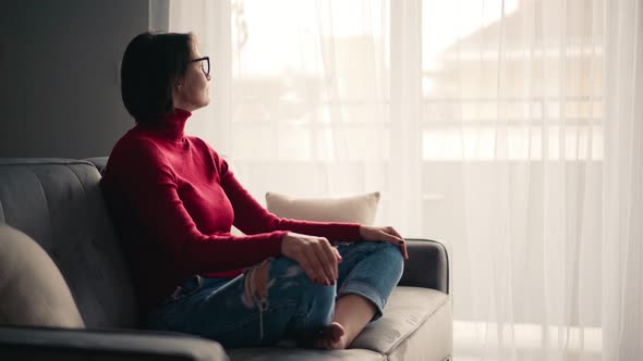 A Young Woman Sitting on the Couch in Her Living Room and Looks Out the Window